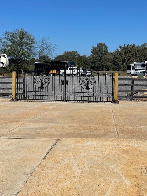 Black metal gate with tree designs stands closed at the entrance to a gravel lot, with parked vehicles and trees visible in the background under a clear blue sky.