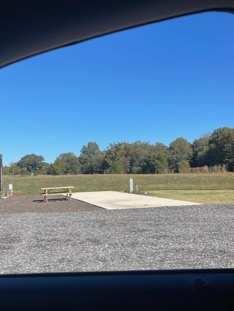 View through a car window of an empty RV campsite with a picnic table, concrete pad, and grassy field under a clear blue sky.