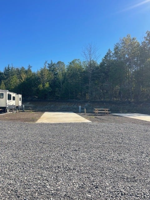 Gravel and concrete RV campsite with utility hookups, a picnic table, and trees in the background under a clear blue sky.